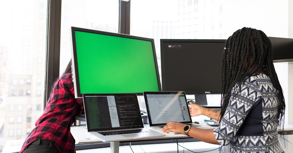 Two women working on laptops and monitors in a bright office setting, focused on technology and teamwork.